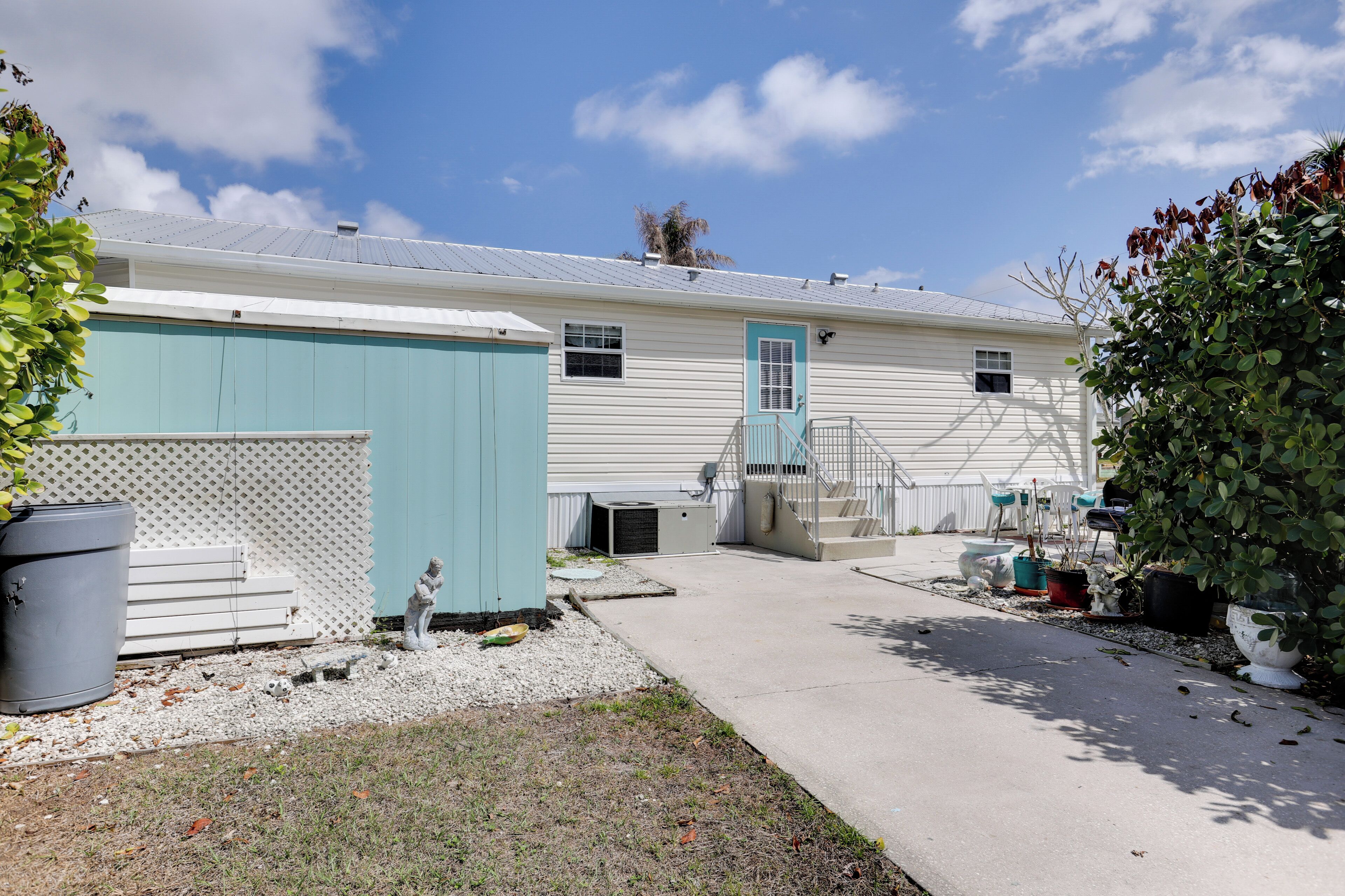 A single-story mobile home with a covered porch, a small front yard with potted plants, and a clear blue sky.