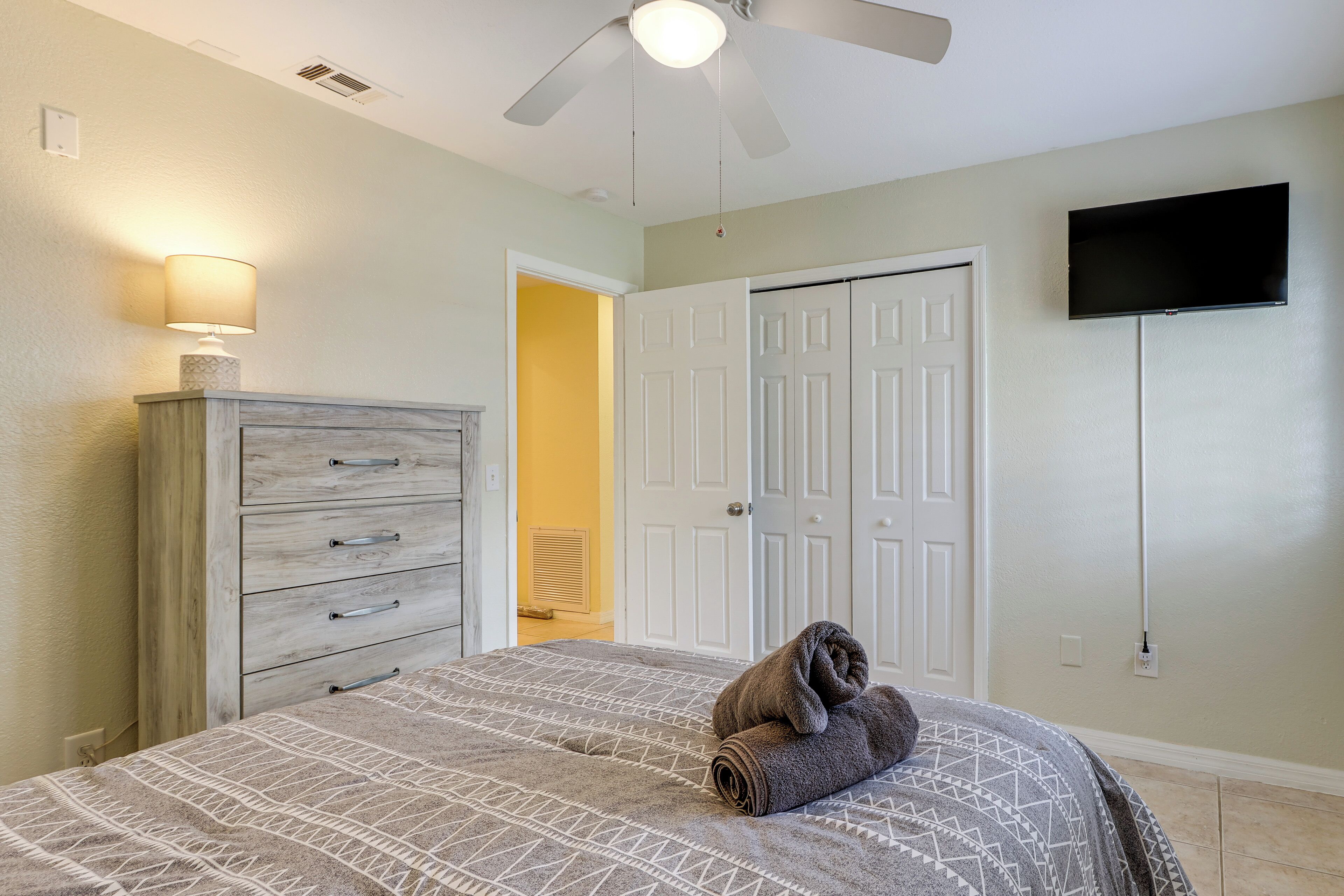 Un dormitorio con cama, cómoda, ventilador de techo, televisor y puerta a otra habitación.