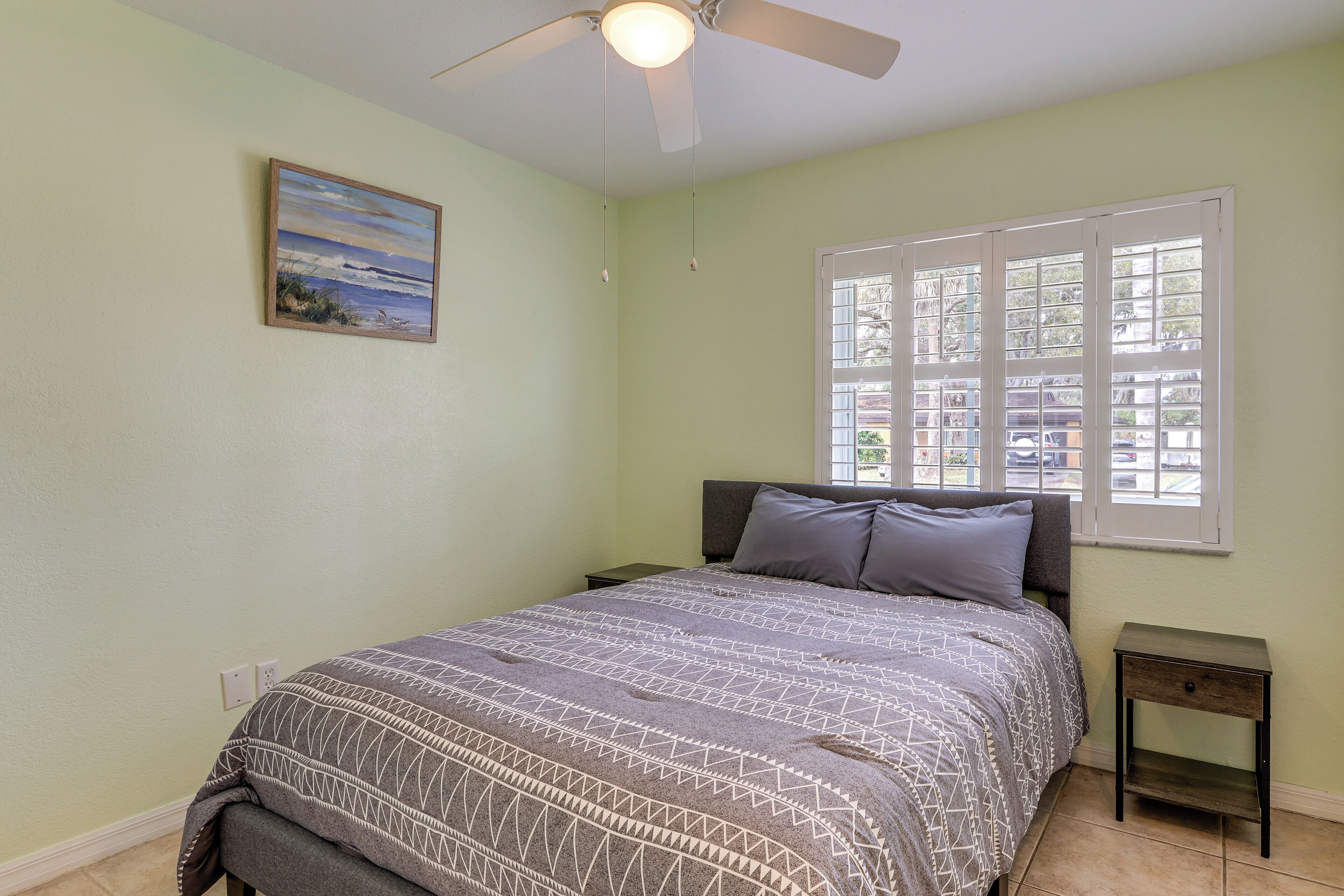 Une chambre à coucher comprenant un lit, une table de chevet, un ventilateur de plafond, une fenêtre avec des stores et un tableau accroché au mur.