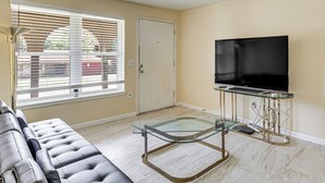 A living room with a glass-top coffee table, a leather sofa, a flat-screen TV, and a door with a window with blinds. - 8 Mi to Bricktown Fun: Quiet Family Retreat (Oklahoma City)