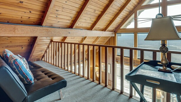 A wooden interior with a sloped ceiling, a cushioned bench with pillows, a table with a lamp, and a view through large windows. - 'point of View' Hilltop Sanctuary in Brevard (Brevard)