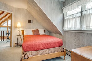 Ein Schlafzimmer mit schrägem Dach, einem Bett mit roter Decke, einem Nachttisch mit Lampe und einem Fenster mit Vorhängen.