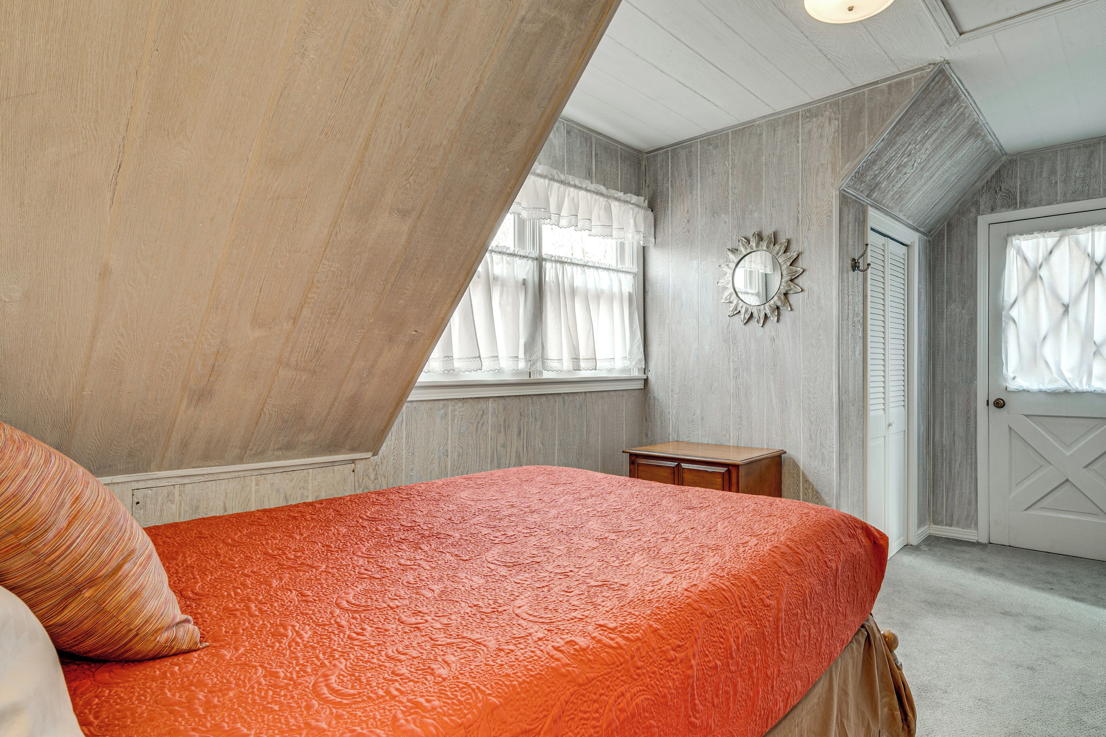 A bedroom with a sloped ceiling, a bed with an orange bedspread, a small wooden nightstand, a mirror on the wall, and a door with a window.
