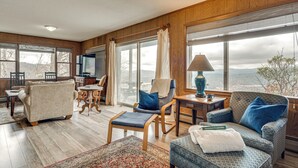 A spacious living room with a large window, wooden walls, and a cozy seating area including a sofa, chairs, and a small table with a lamp. - 'point of View' Hilltop Sanctuary in Brevard (Brevard)