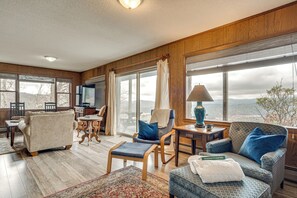 A spacious living room with a large window, wooden walls, and a cozy seating area including a sofa, chairs, and a small table with a lamp. - 'point of View' Hilltop Sanctuary in Brevard (Brevard)