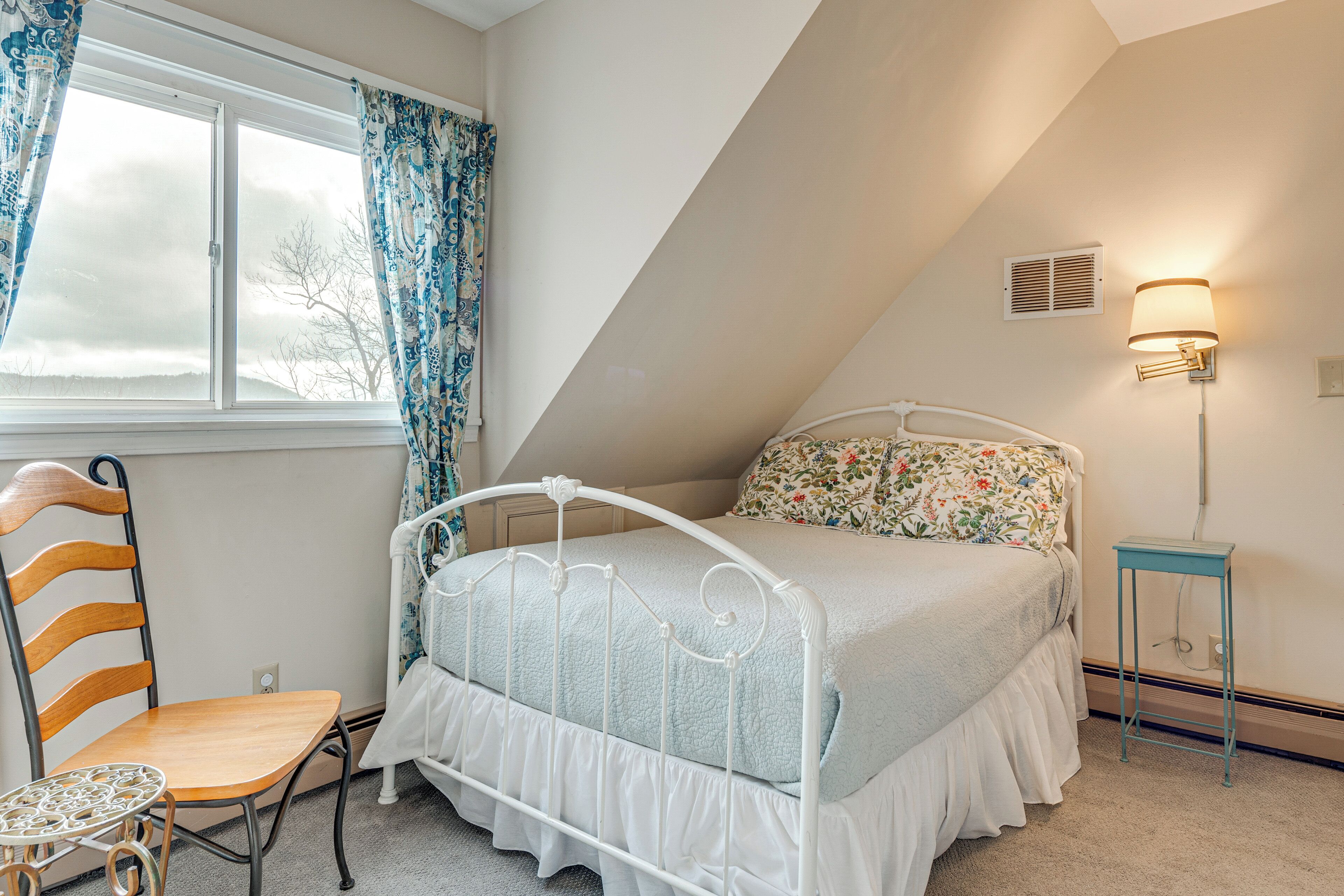 Ein Schlafzimmer mit einem Bett, einem Sessel, einem kleinen Tisch, einem Fenster mit Vorhängen und einer Wandlampe.