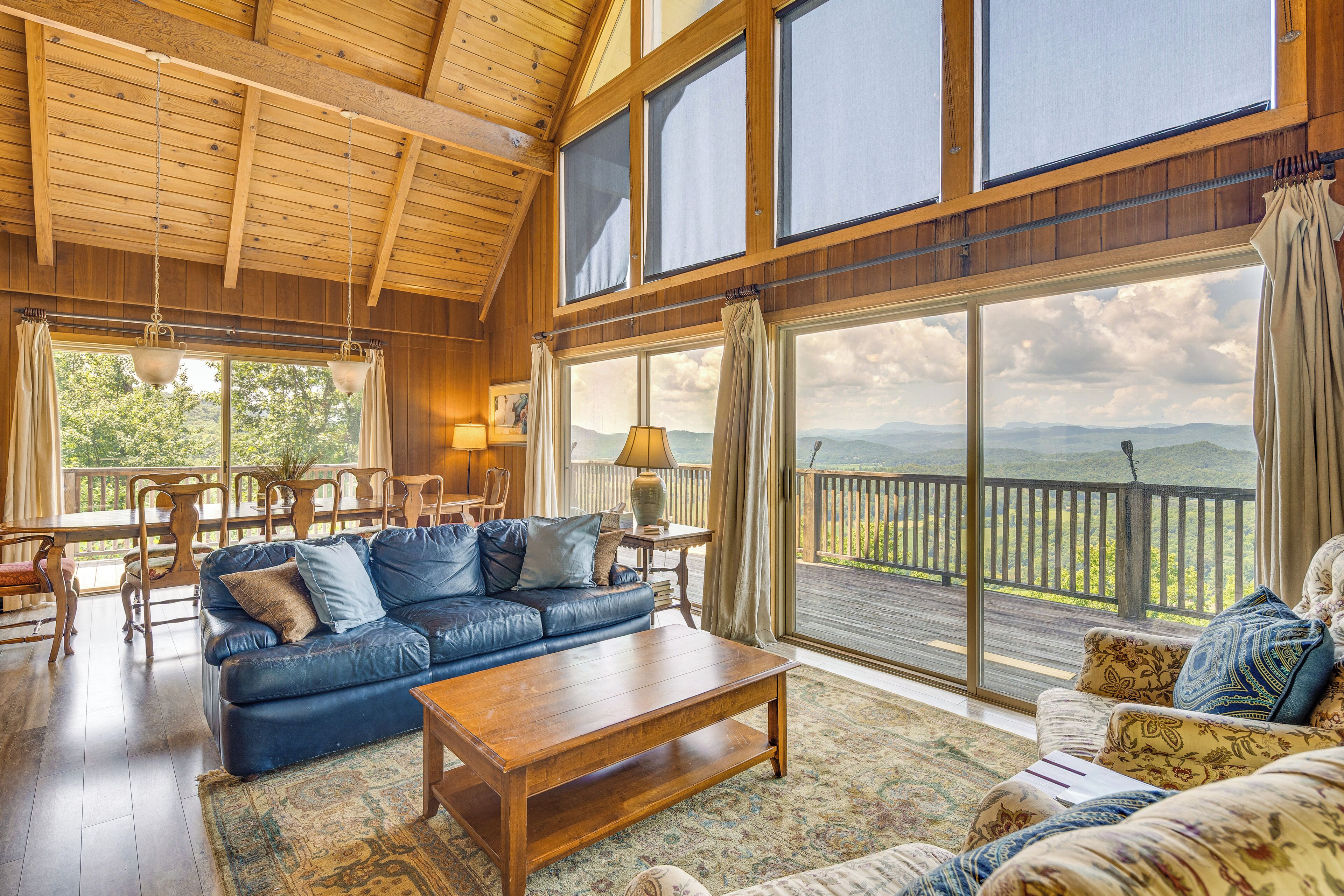 A spacious living room with a large wooden ceiling, a blue leather sofa, a wooden coffee table, and a view of a mountainous landscape through large windows.