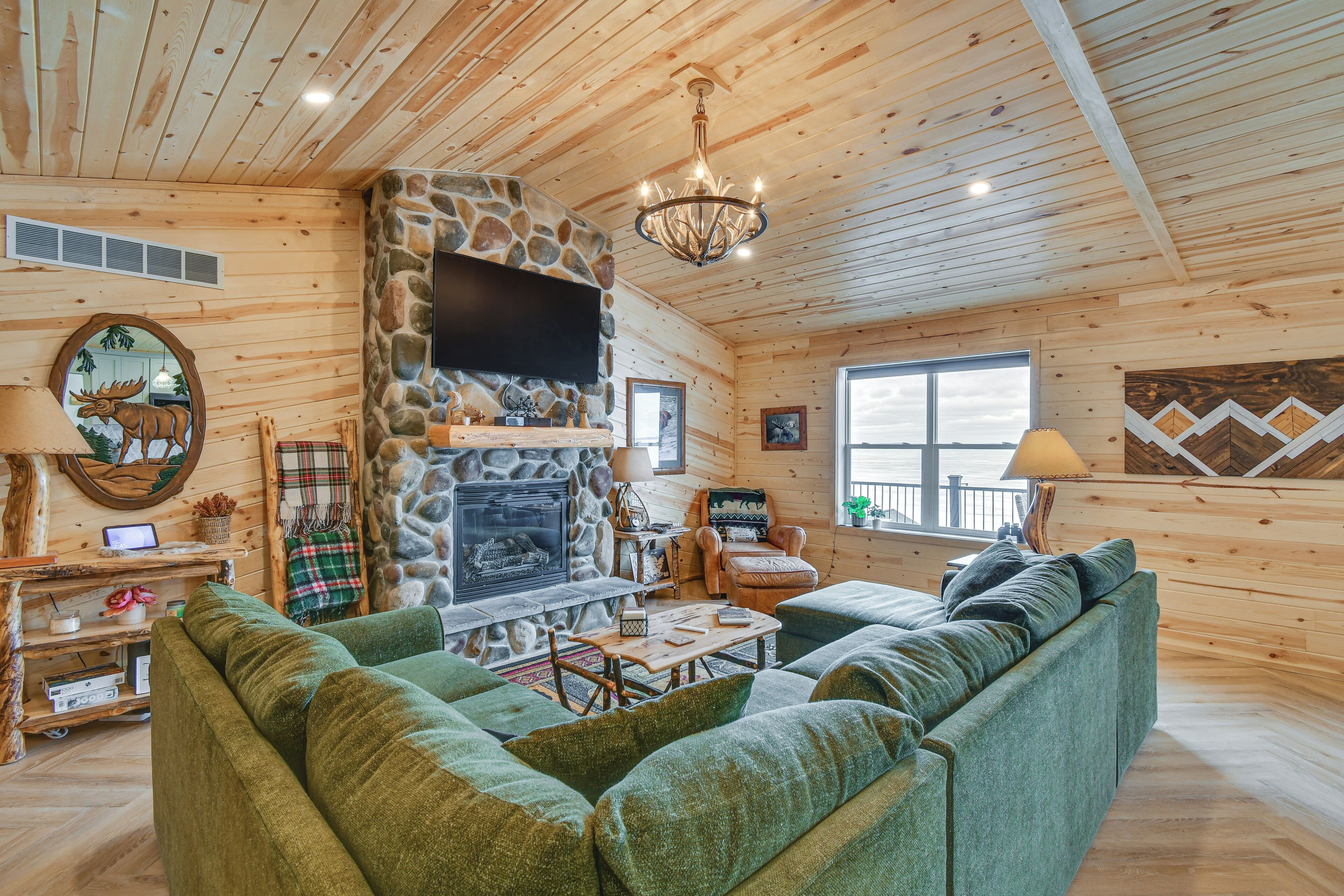 A cozy log cabin interior with a stone fireplace, green upholstered sofa, and a television mounted on the wall.