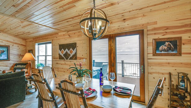 A wooden cabin interior with a dining table, chairs, a chandelier, and a large window. - Luxe Hideaway on Houghton Lake: Families Welcome! (Houghton Lake)