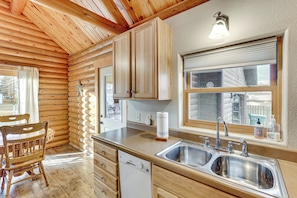 Interieur van een houten blokhut met een keuken, inclusief een dubbele spoelbak, houten kastjes, een eettafel met stoelen en een raam met uitzicht op een witte schutting.