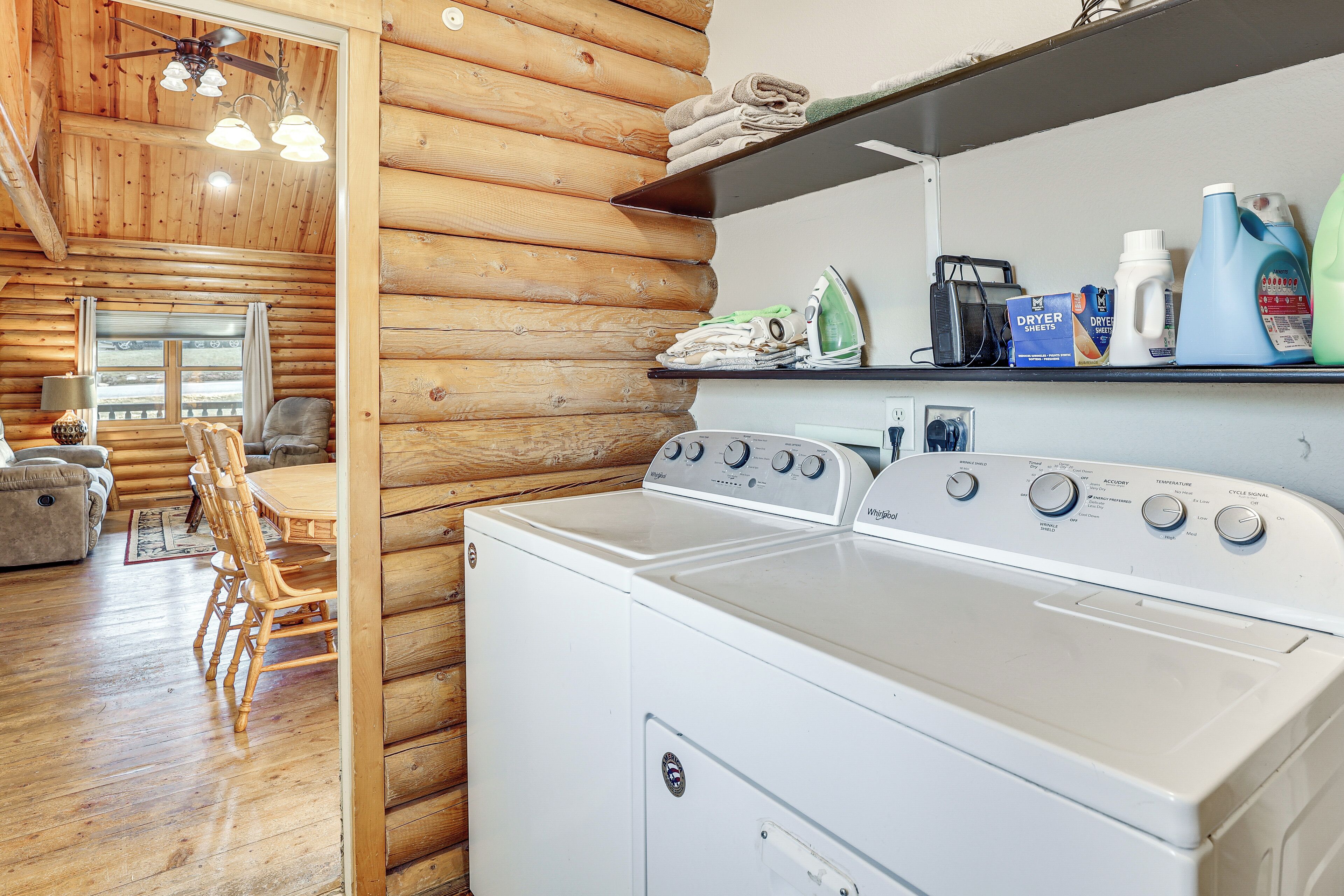A laundry area with a front-loading washer and dryer, a wooden dining table with chairs, and a shelf with laundry supplies.