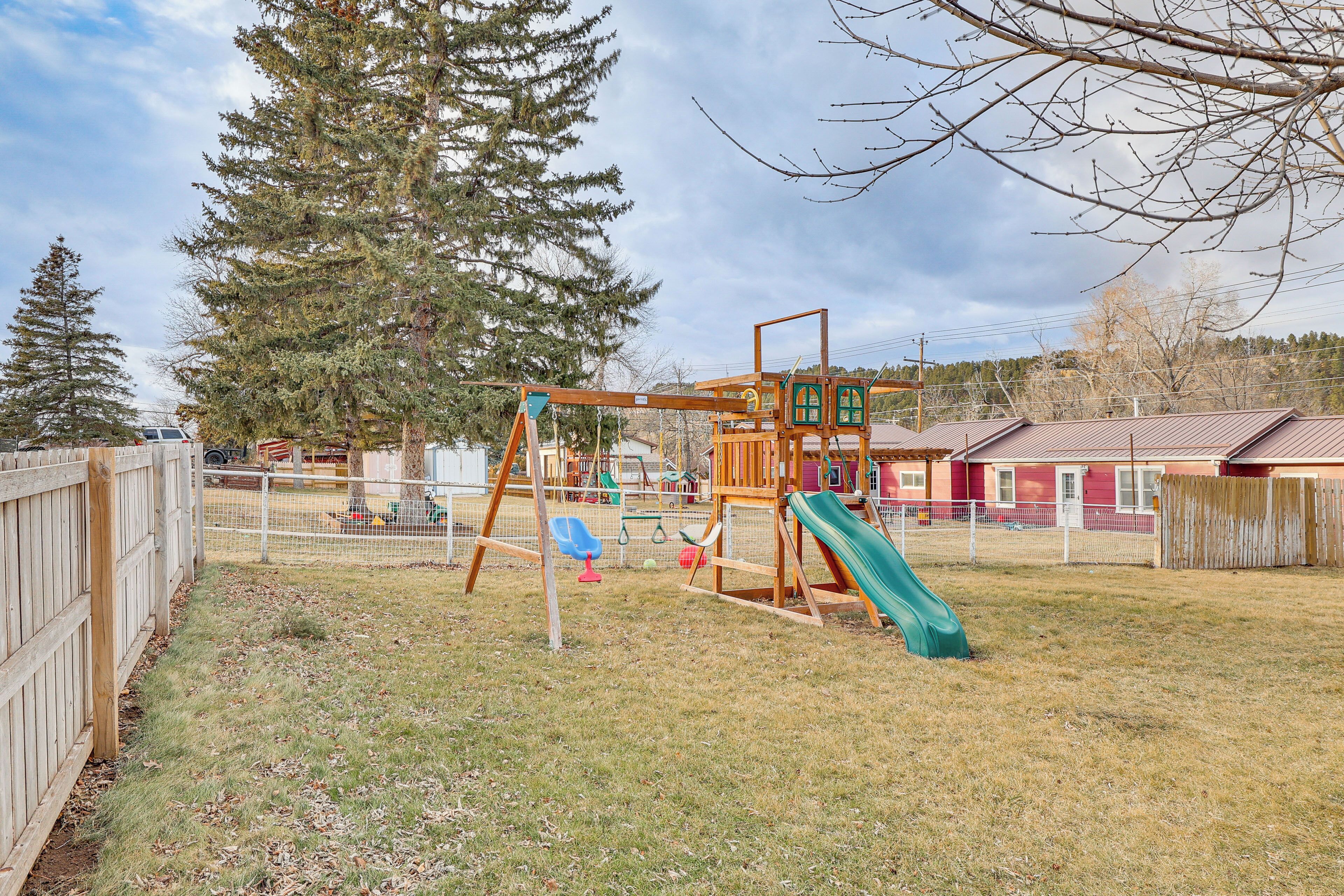 A playground with a slide and swings, surrounded by a wooden fence and trees.