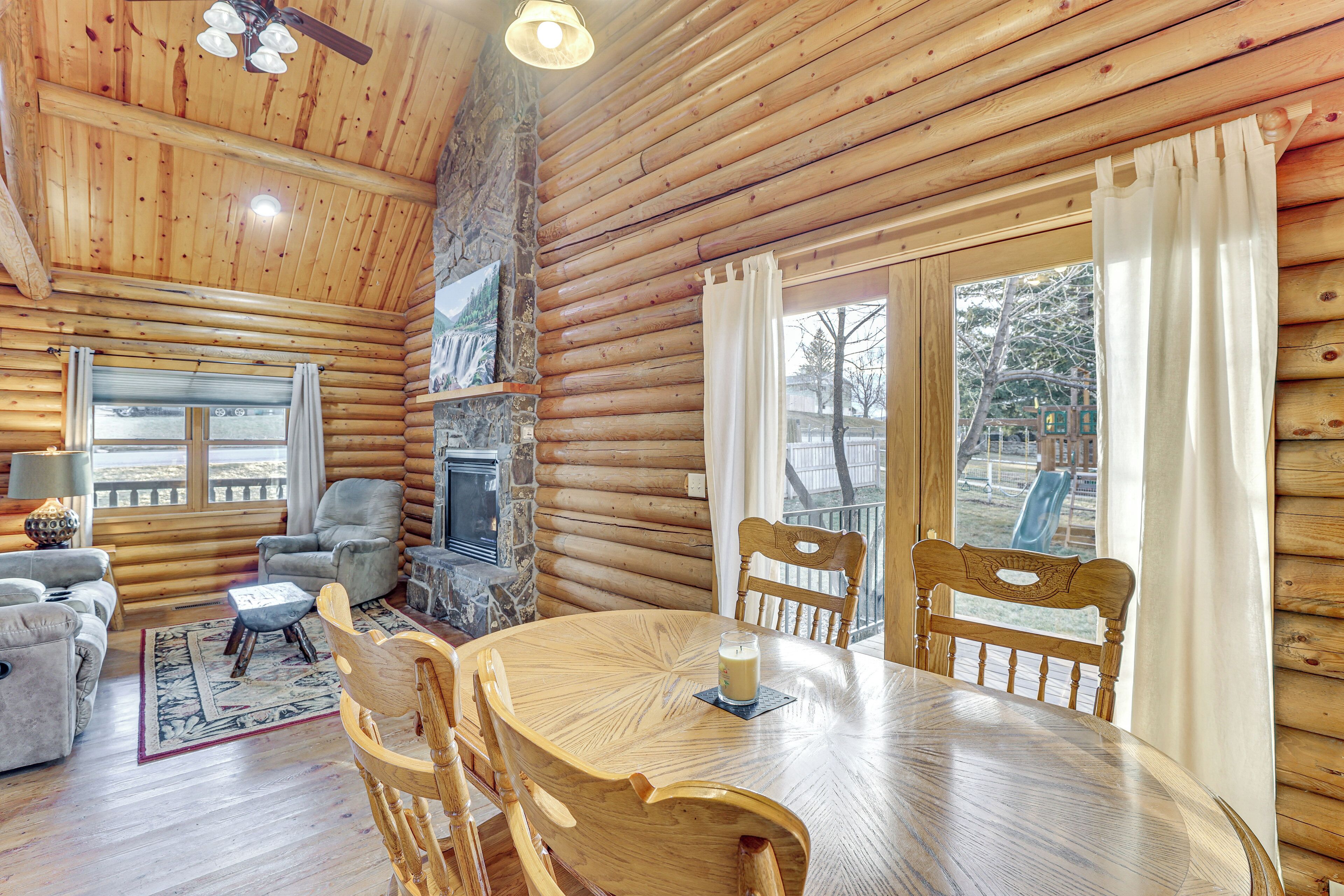 A log cabin interior with a round wooden dining table, two wooden chairs, a fireplace, and a stone wall.