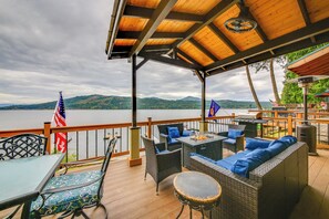 A covered outdoor deck with wicker furniture, a table, and chairs, overlooking a body of water and hills. - Hike, Fish & Unwind: Cocolalla Lake Home w/ Dock! (Cocolalla)