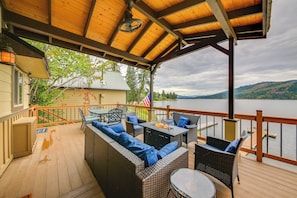 A covered deck with wicker furniture, a fire pit, and a view of a lake and mountains. - Hike, Fish & Unwind: Cocolalla Lake Home w/ Dock! (Cocolalla)