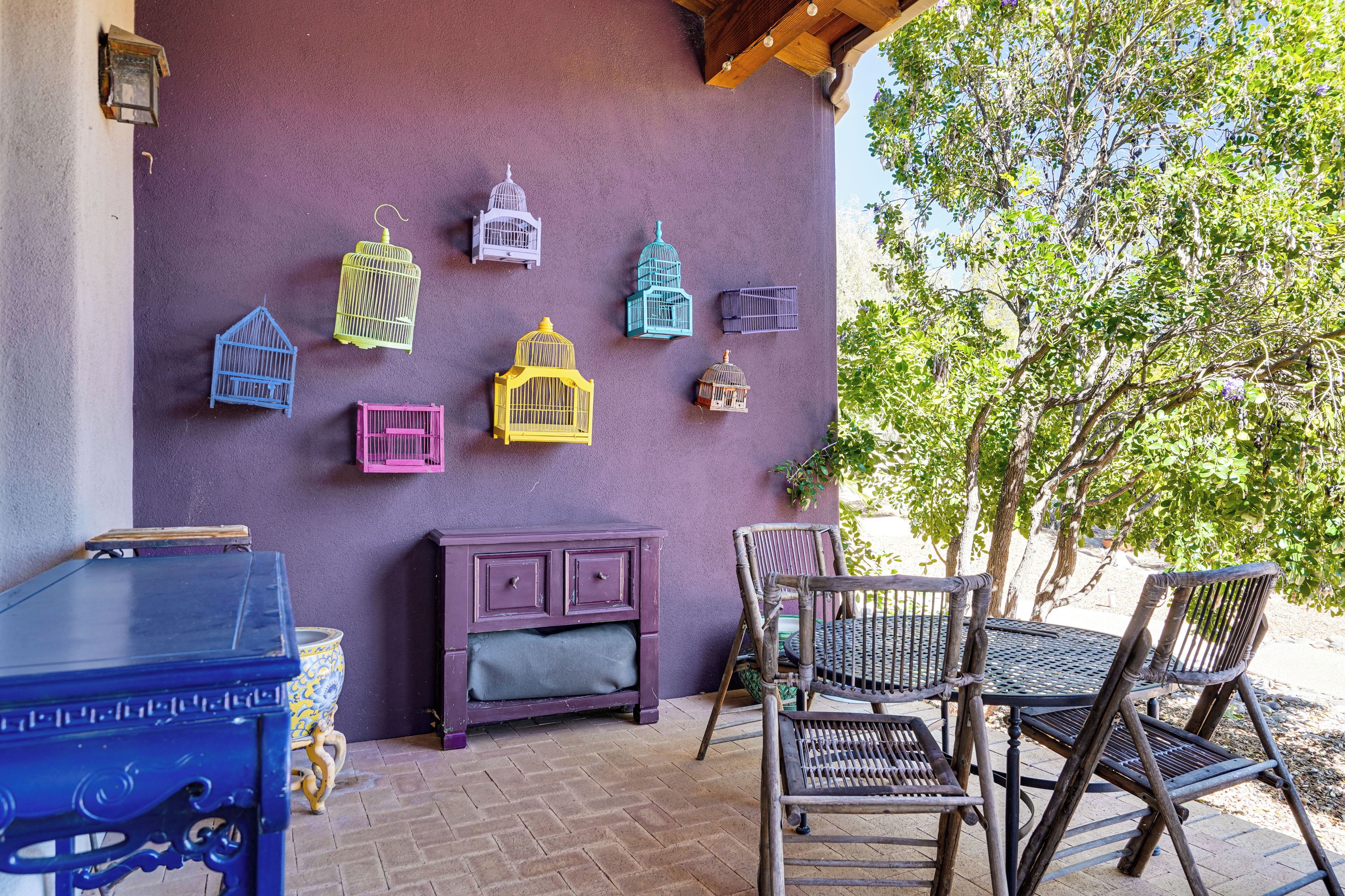 Un patio con un muro morado, sillas de madera, una mesa y decoraciones de jaulas de pájaros.
