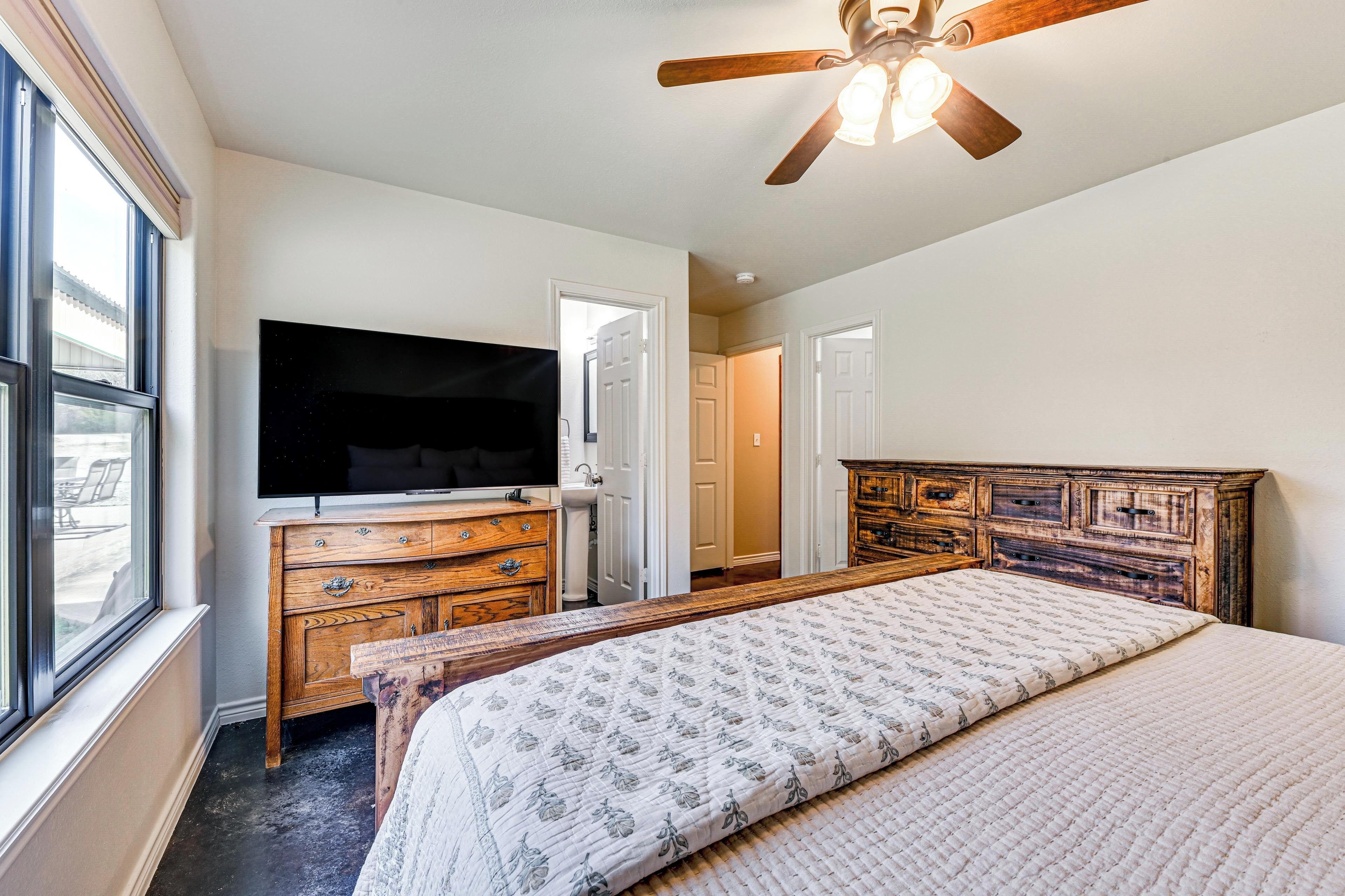 A bedroom with a bed, a TV, a wooden dresser, a ceiling fan, and a window.