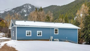 Une maison bleue d’un seul étage, avec un toit recouvert de neige, une allée en gravier et une montagne boisée en arrière-plan.