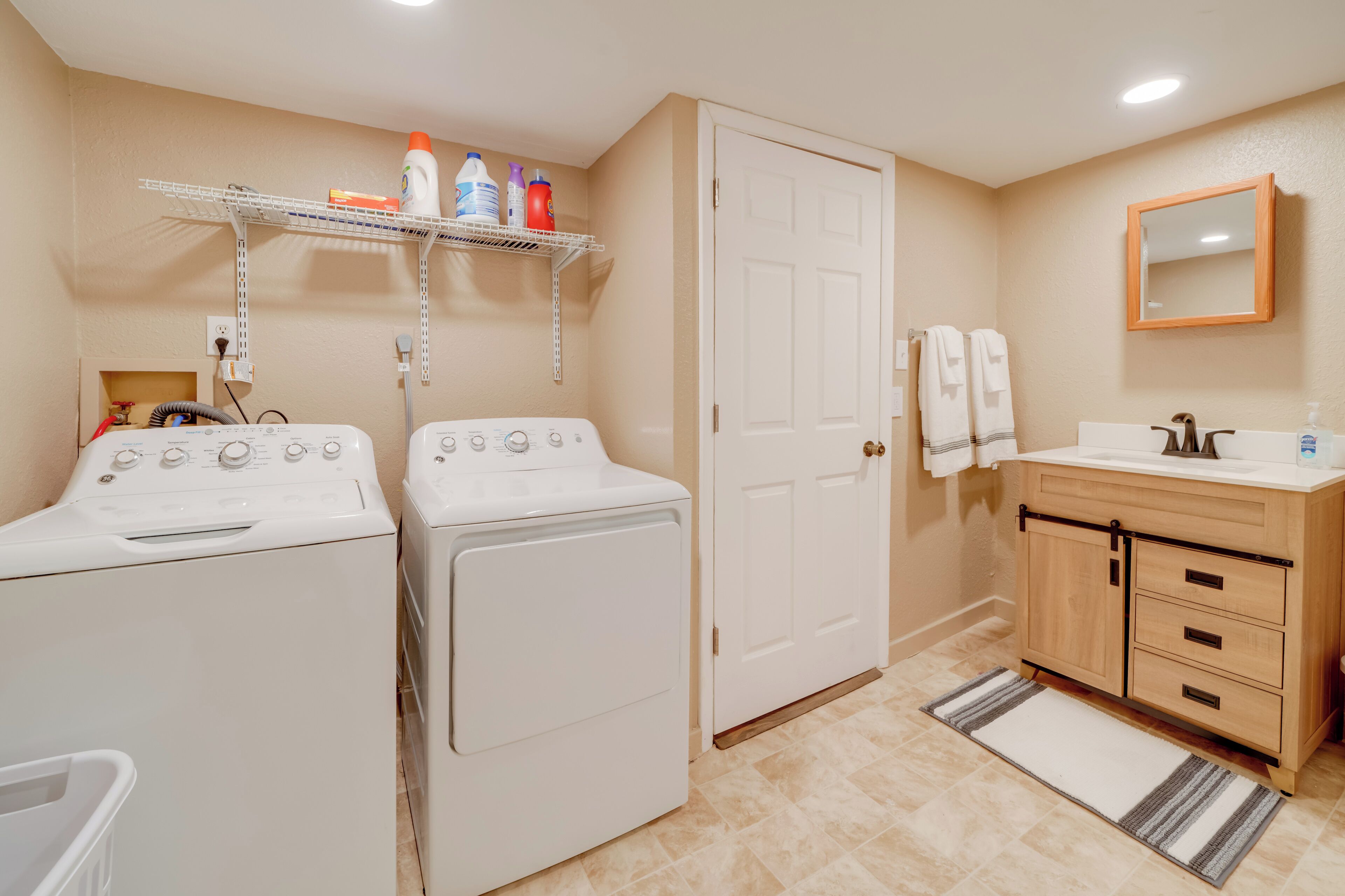 Une buanderie équipée de deux machines à laver à chargement frontal, d’une étagère contenant des produits de nettoyage, d’une porte blanche, d’un meuble-lavabo en bois avec un miroir et d’un tapis à rayures.