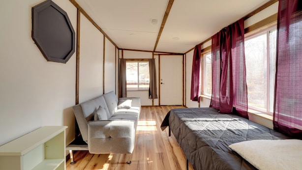 A modern room with a wooden floor, a grey sofa, a bed with a grey duvet, and a window with red curtains. - 4 Mi to Gauley River Rapids! Outdoor Adventure Hub (Summersville)