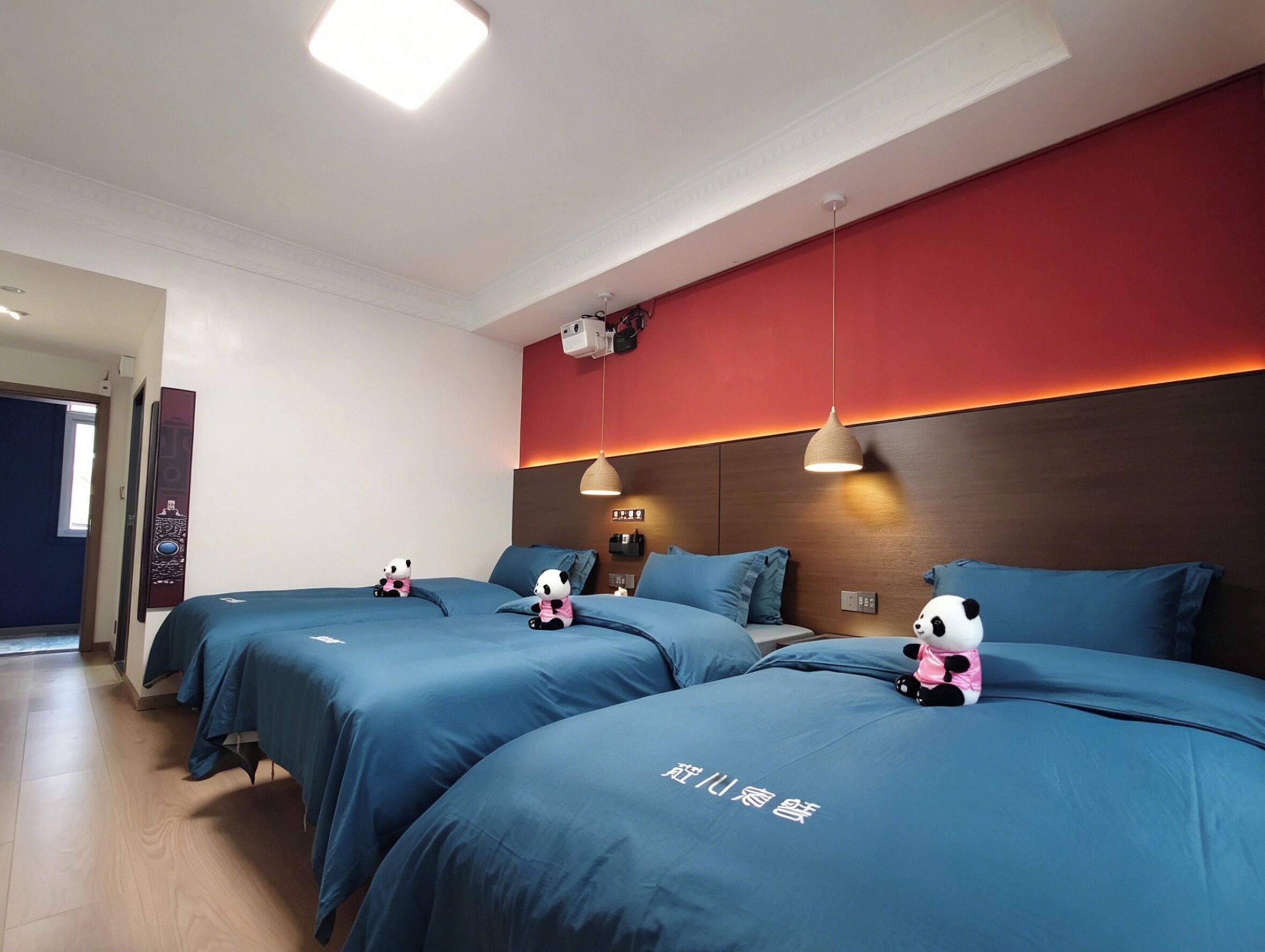 A hotel room with three beds, each with a blue bedspread and a panda plush toy. There is a red accent wall, a ceiling-mounted projector, and a wooden headboard.