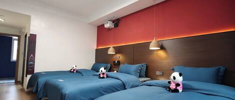 A hotel room with three beds, each with a blue bedspread and a panda plush toy. There is a red accent wall, a ceiling-mounted projector, and a wooden headboard.