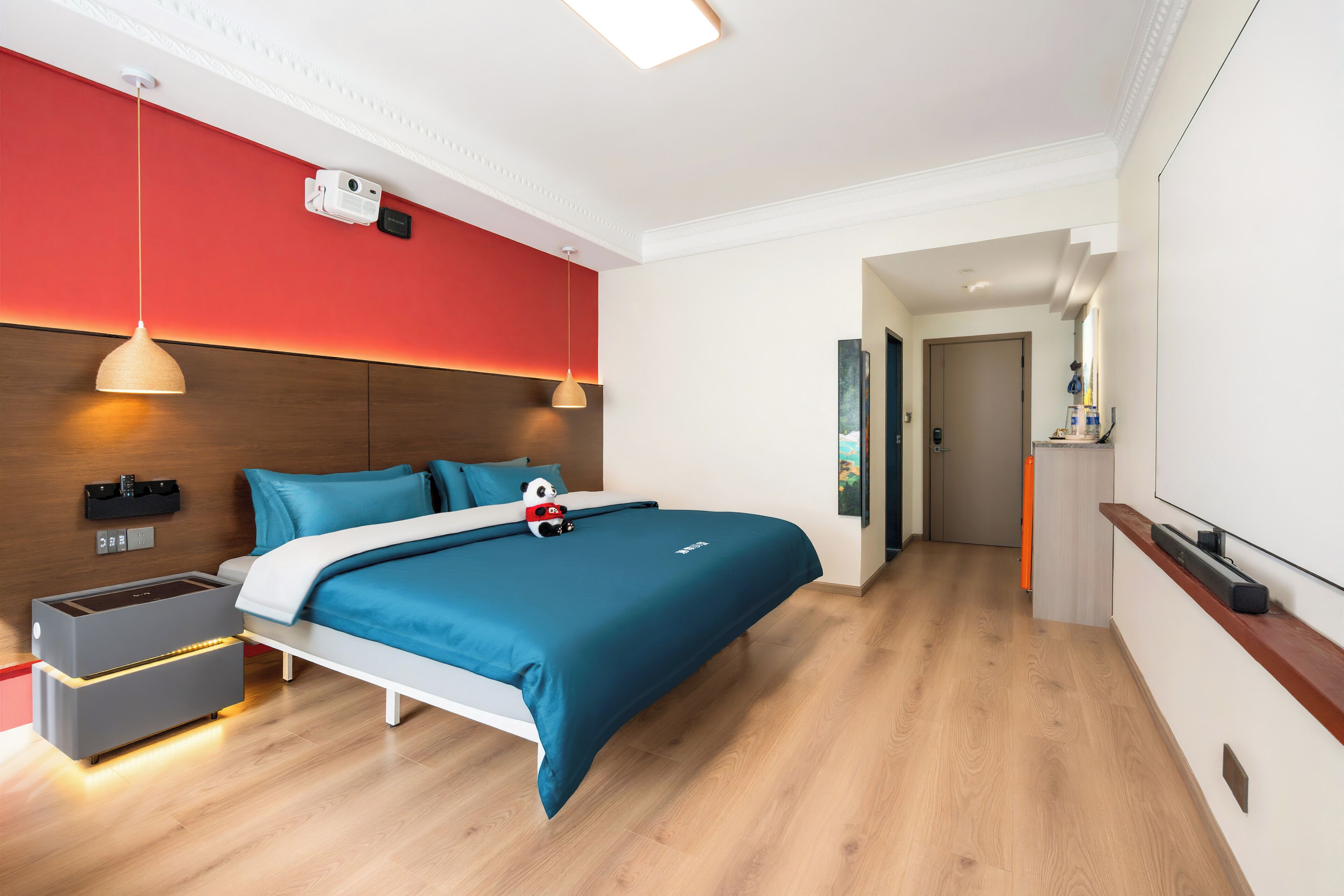 A modern hotel room with a large bed, a bedside table, a nightstand, a red accent wall, and a wooden floor.