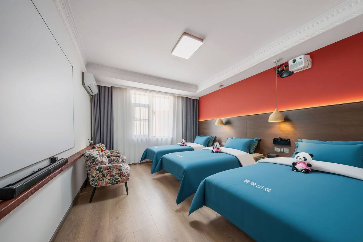 A hotel room with three beds, a wooden floor, a large window with curtains, a ceiling-mounted projector, and a panda plush toy on one bed.