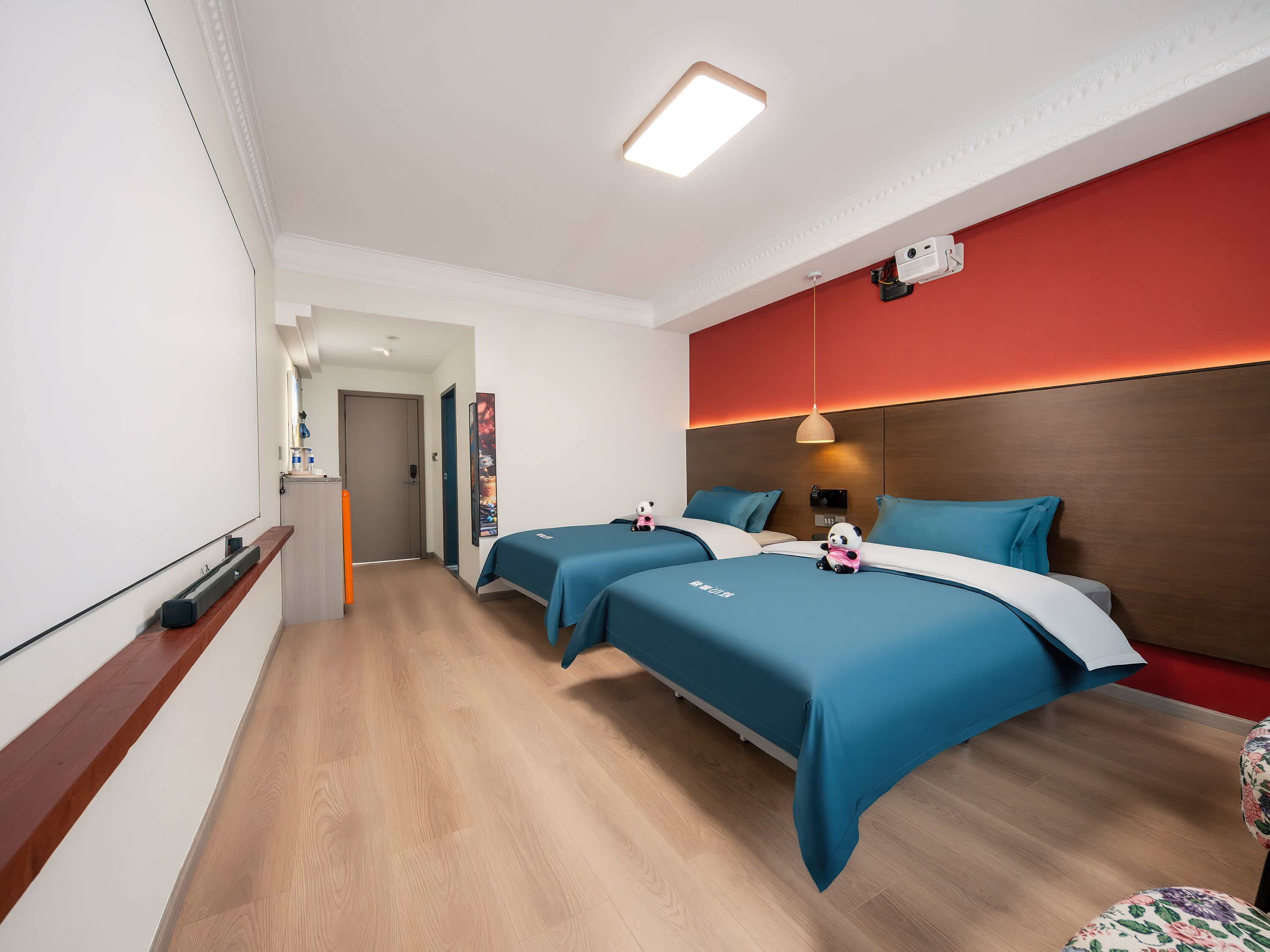 A hotel room with two beds, a red accent wall, a wooden headboard, and a ceiling-mounted projector.