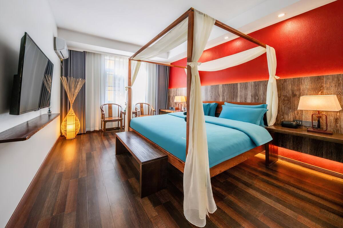 A four-poster bed with a canopy, a television on the wall, a bedside table with a lamp, and a wooden bench.