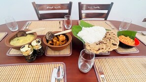 Pyöreä ruokapöytä, jonka päällä on erilaisia ruokia, kuten riisiä, leipää ja curryruokia, ja pöydällä on kudottu alusta.