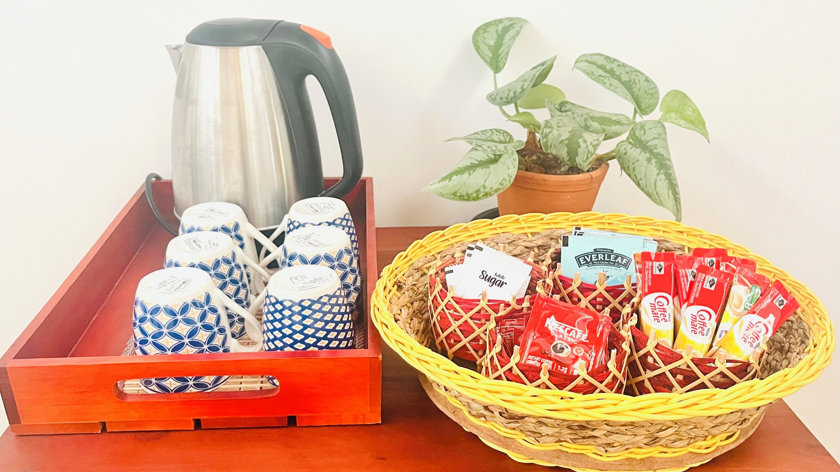 Un plateau avec une bouilloire et des tasses à motifs, un panier contenant des sachets de café instantané et une plante en pot.