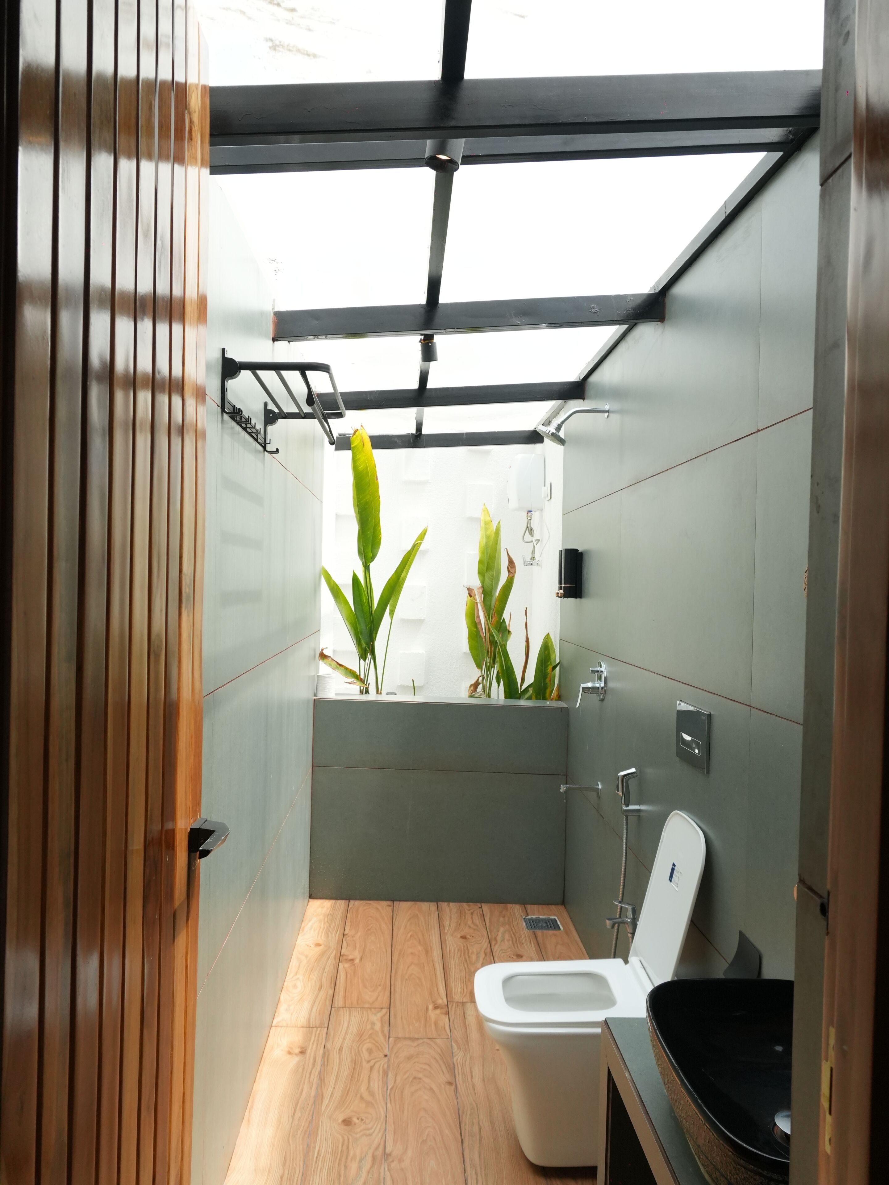 A modern bathroom with a wooden floor, a white toilet, a black sink, and a window with plants.