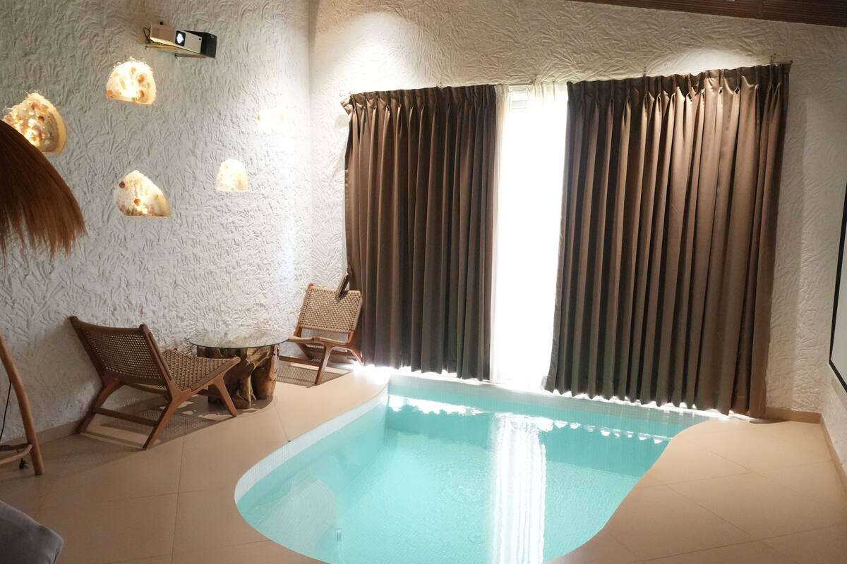 A swimming pool with a curved edge, a chair, a table, and a wall with textured plaster and wall-mounted lights.