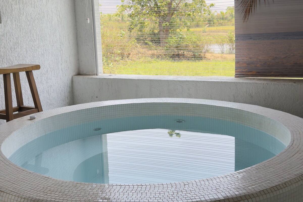 A circular hot tub with tiled walls, a wooden bench, and a view of a tree and landscape through a window.