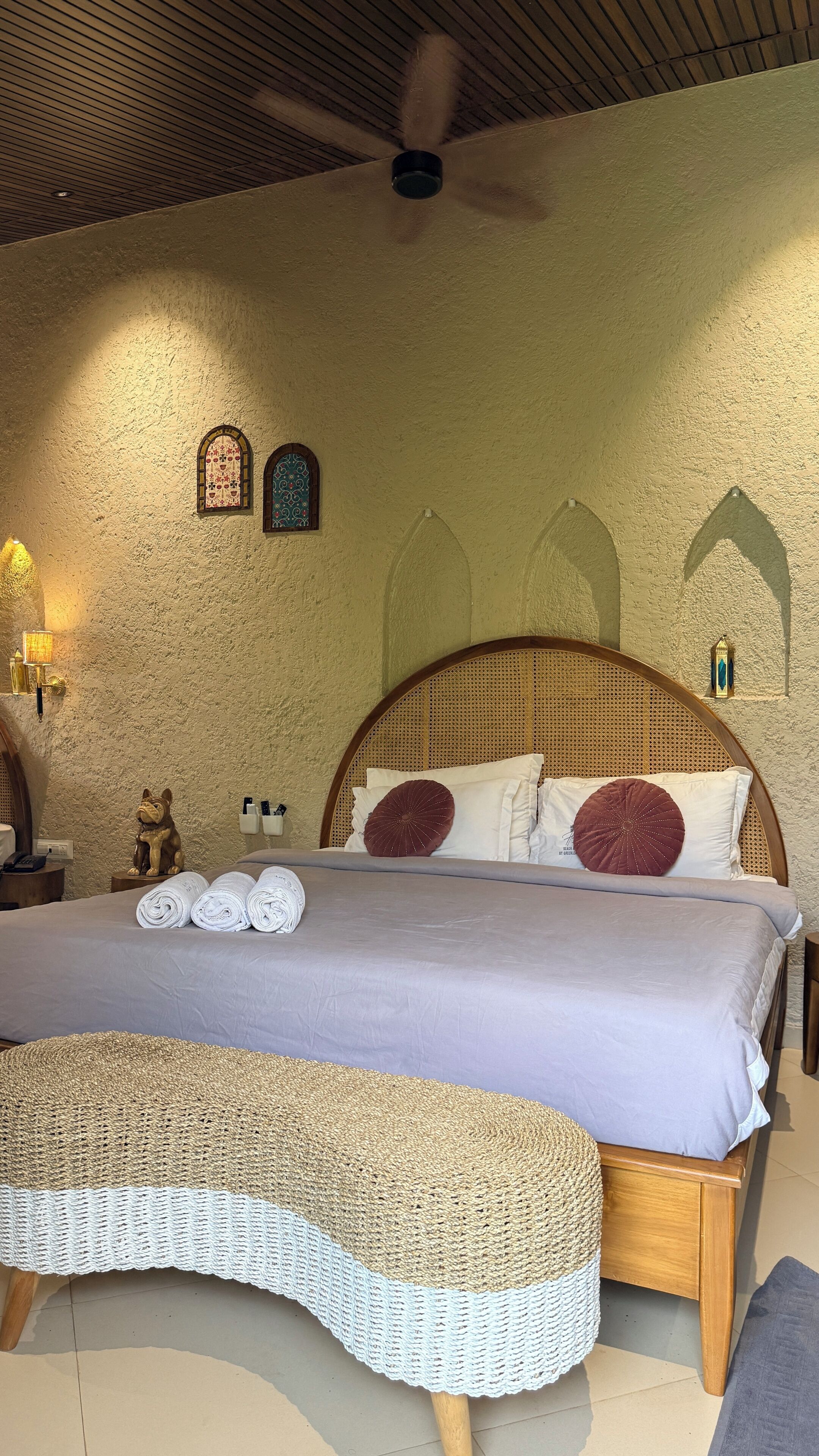 Ein Schlafzimmer mit einem hölzernen Bett, einer gewobenen Bank, einem Deckenventilator und zwei Wandleuchten.