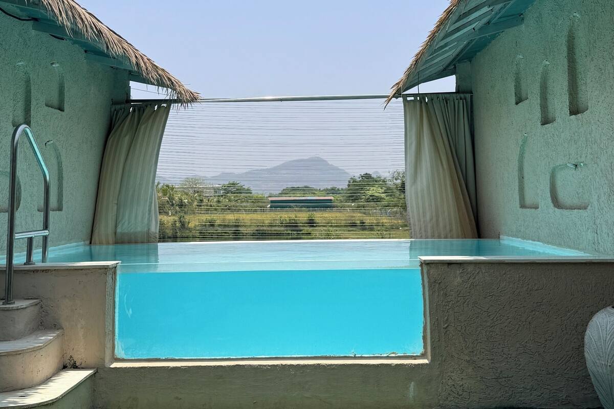 A swimming pool with a view of a mountain, surrounded by a building with a thatched roof and a green lawn.