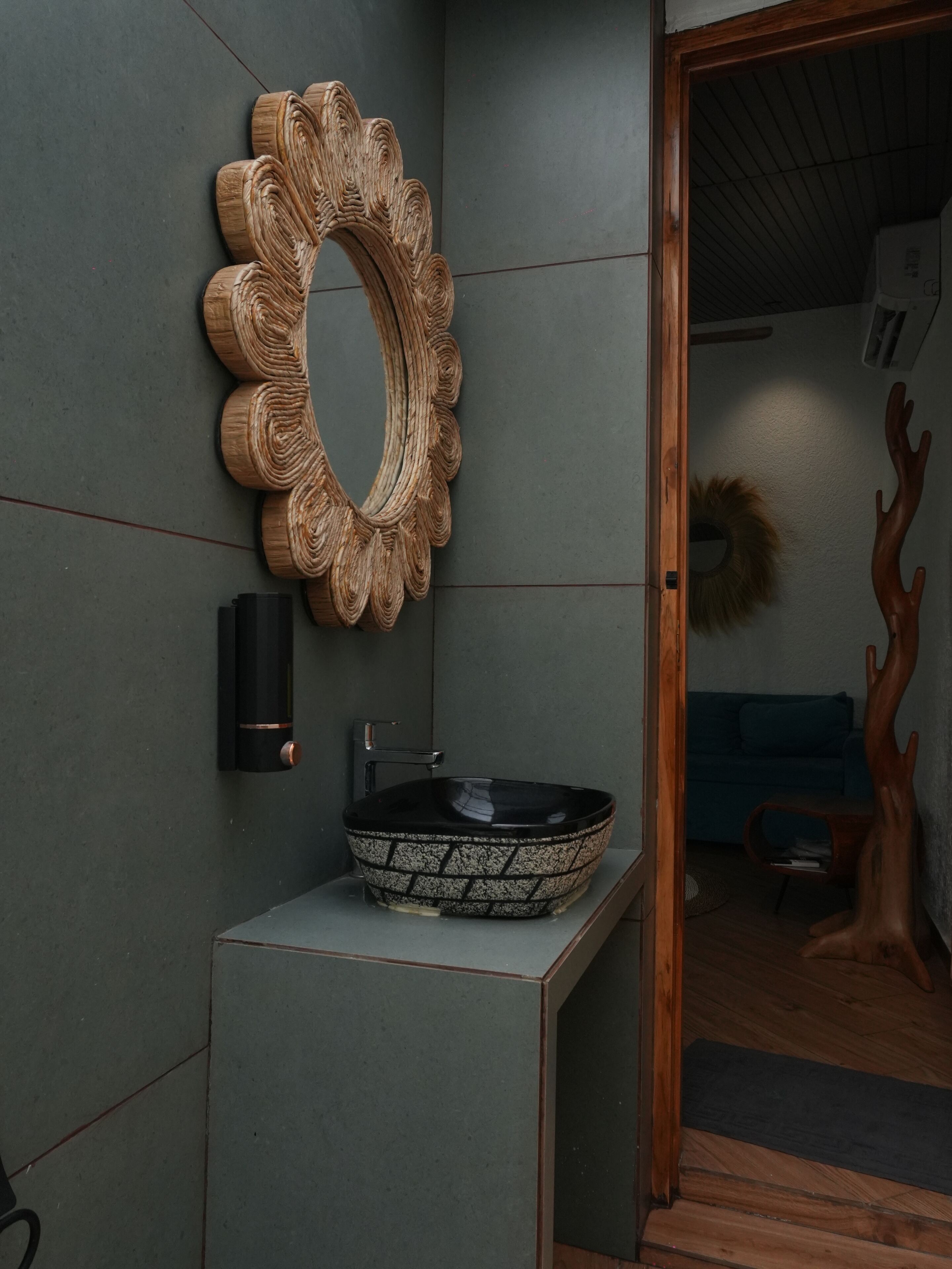 Un baño con espejo de marco de madera, un lavabo tipo cuenco negro y un adorno con forma de árbol de madera.