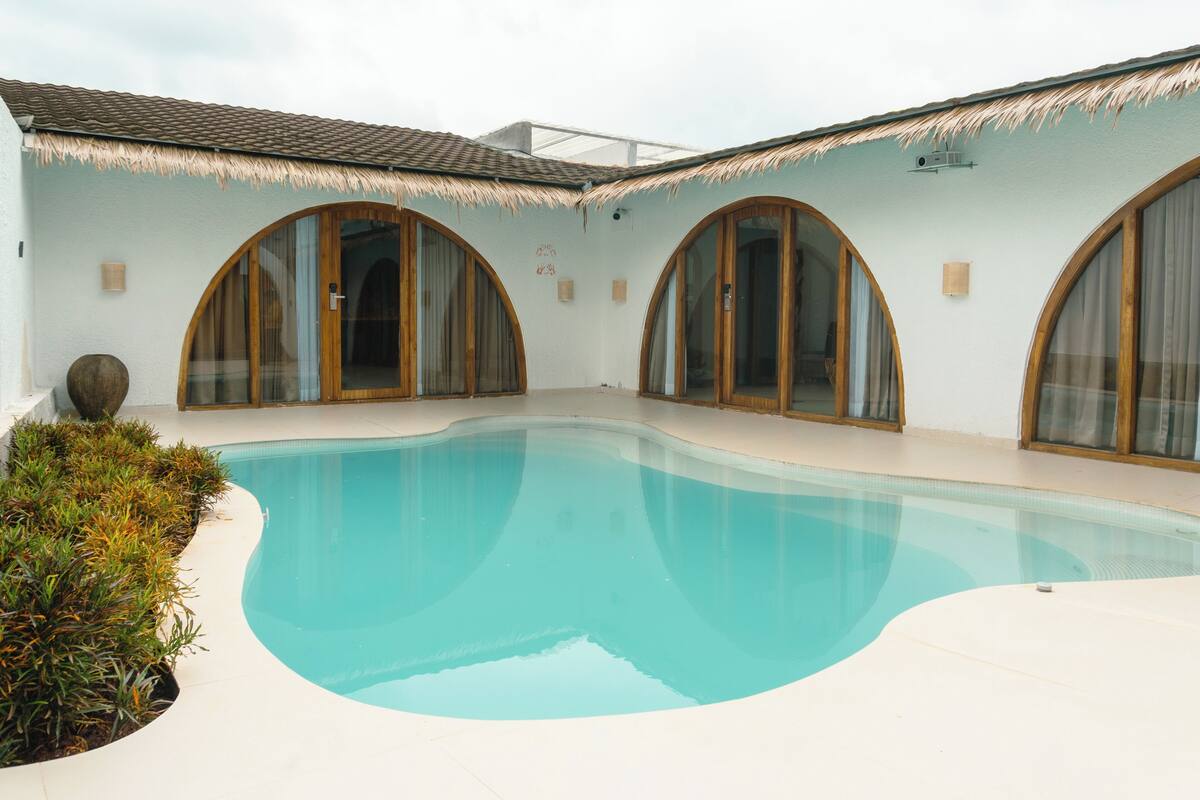A pool area with a curved design, large arched windows, and a thatched roof.