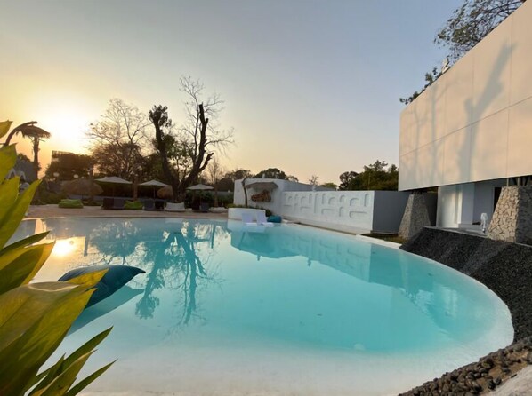 A swimming pool with lounge chairs, a white wall with a textured design, and trees in the background. - Hidden Beach Resort by Greenleaf Aqua (Karjat)