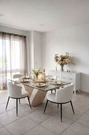 Un espace repas moderne avec une table en verre, des chaises blanches et une console blanche ornée d’un vase de fleurs.