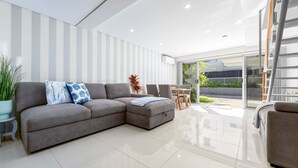 A modern living room with a grey sectional sofa, a small table, and chairs. There is a sliding glass door leading to an outdoor area. - The Wharfside Oasis by the Waves (Surfers Paradise)