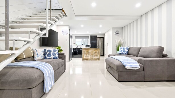 A modern living room with a grey sofa, a blue-striped throw blanket, a staircase, a kitchen area, and a wall-mounted TV. - The Wharfside Oasis by the Waves (Surfers Paradise)