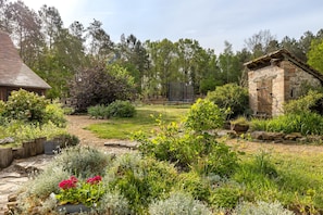 Un chalet rustique aux murs de pierre, entouré d’un jardin bien entretenu où poussent différentes espèces de plantes et délimité par une clôture en bois.