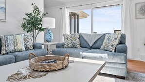 A modern living room with a grey sofa set, a coffee table, and a large window offering a view of the sea.