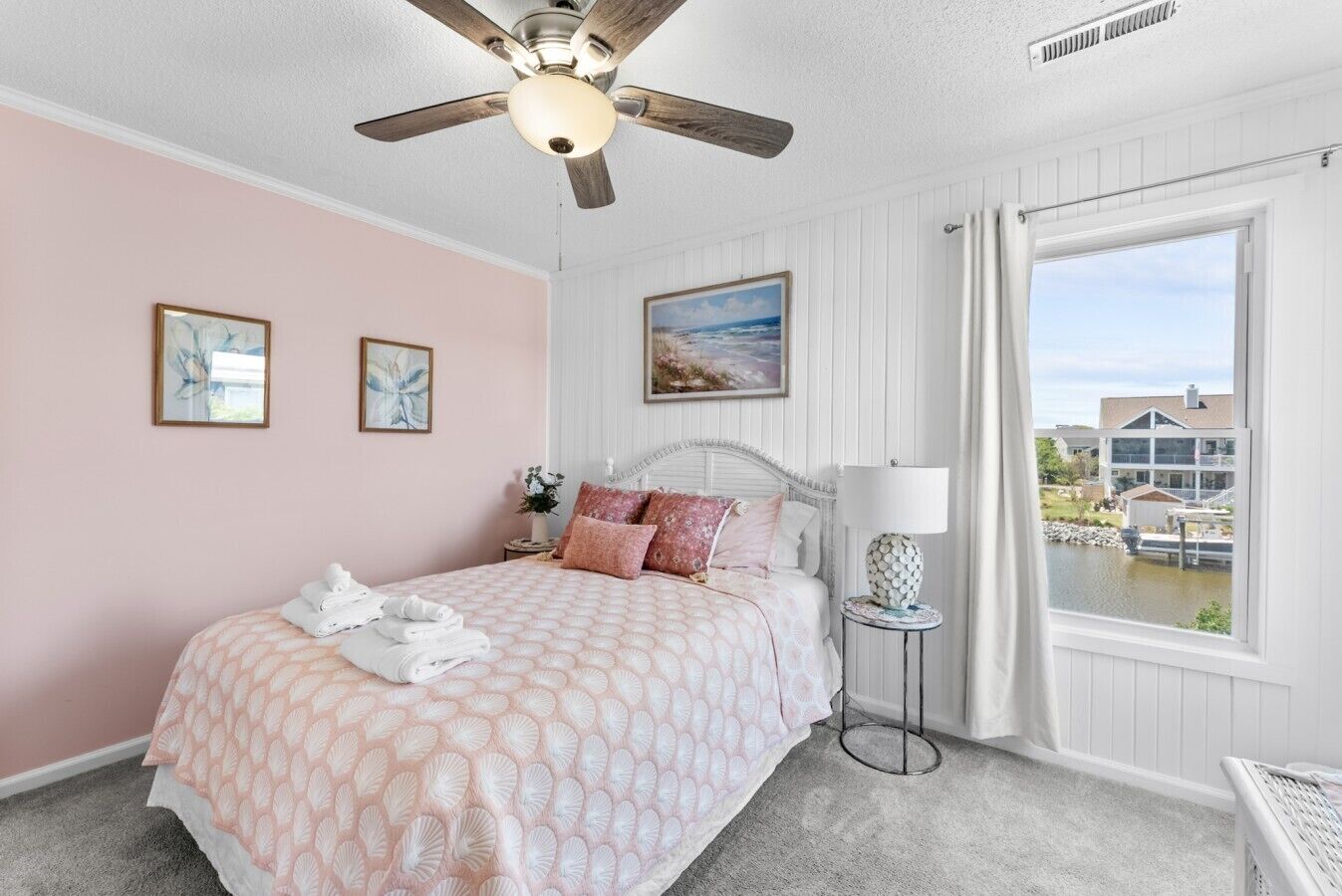 A bedroom with a bed, a ceiling fan, a window with a view, and a small table with a lamp.