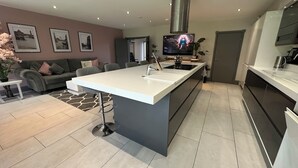 A modern kitchen with a central island, a dining area with a sofa, and a TV mounted above the island. - Luxury 5-Bed Hale Home | Cinema, Pool Table & Sleeps 10 (Hale Barns)