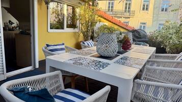 Un patio exterior con mobiliario de mimbre, una mesa blanca con baldosas estampadas y plantas en macetas.