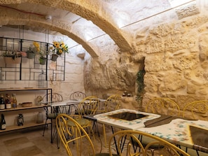 A rustic interior with stone walls, a wooden bar, yellow metal chairs, and a table with a floral tablecloth.