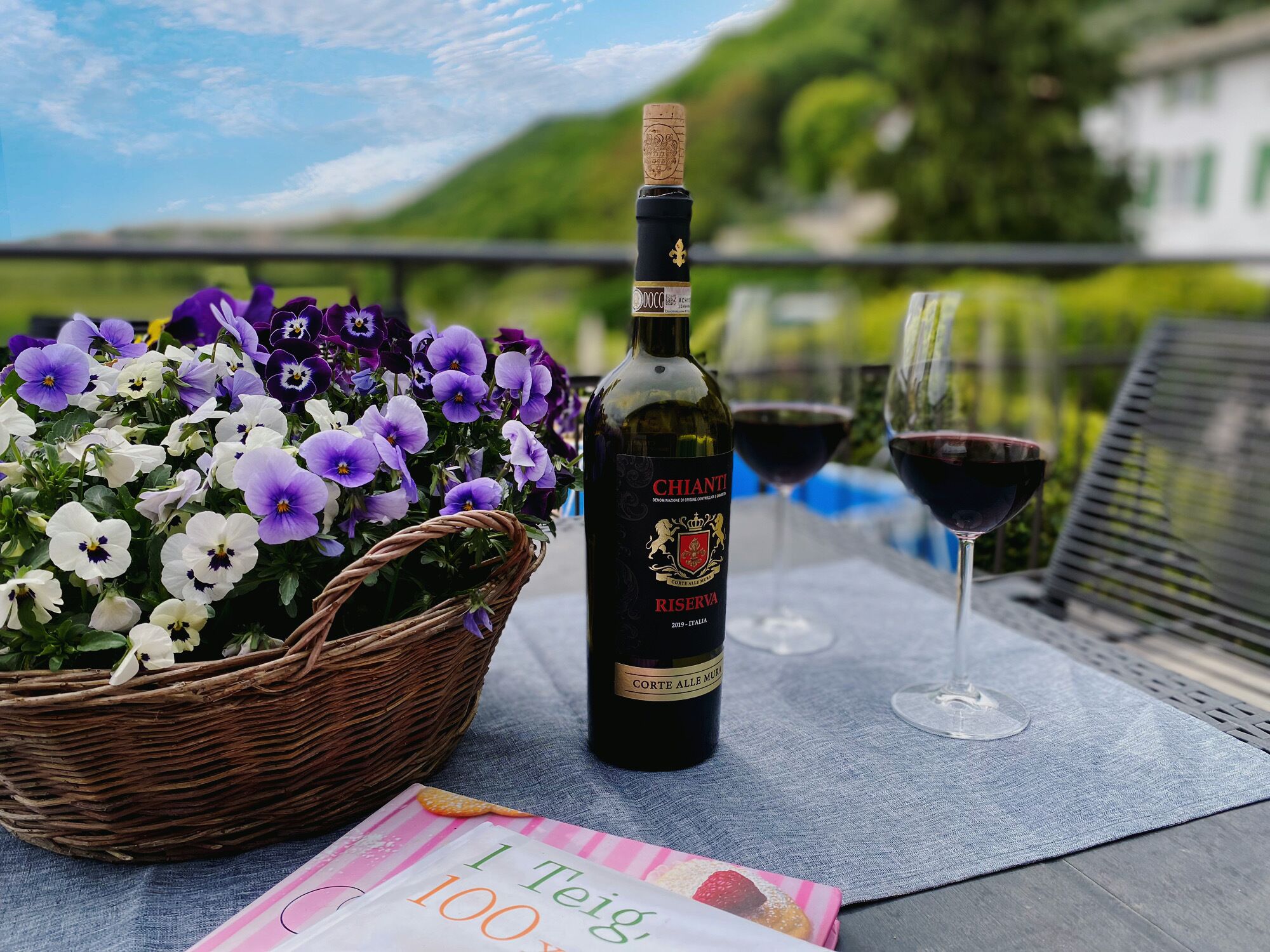 Una botella de vino Chianti Riserva, una cesta de flores moradas y blancas, y dos copas de vino sobre una mesa con una vista panorámica de fondo.